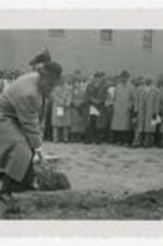 Man shovels dirt at a groundbreaking ceremony with spectators in background.