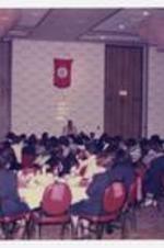 Men and women sit at dining tables in the audience while a man stands at a podium "Oak Brook, Marriot Hotel", in a large banquet room.