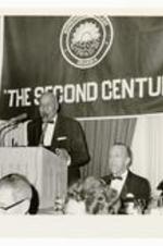 View of a man at a podium with microphone, banner in background "The Second Century."