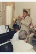 Wynton Marsalis plays his trumpet with a man playing the piano, in a dining room while men and women sit at dining tables.