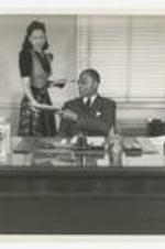 James P. Brawley sits with two other men at a desk while a woman hands him papers.