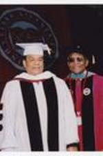 Andrew Young and Walter Broadnax, wearing graduation cap and gowns, stand next to the podium on stage at commencement.