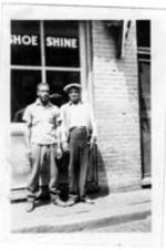 Two unidentified men stand on a sidewalk in front of a building window reading "shoe shine."