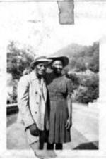 An unidentified couple stand with their arms around each other. They are surrounded by trees on a sunny day.