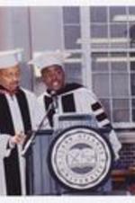 Two men, wearing graduation cap and gowns, stand at the podium at commencement.
