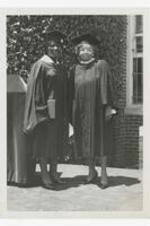 Addie Shropshire and Laura Nesbitt Thompson, wearing graduation caps and gowns, pose in front of a building.