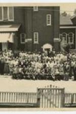 Outdoor group portrait of men, women and children in front of a brick building. Written on verso: 1st row left to right, 5th - James H. Touchstone, 6th - Mrs. Dessie Touchstone.