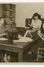Three women pose at a long table with typewriters and papers. Written on verso: CC 1951, Panther Newspaper Staff, R to L - Juanita Traylor - staff typist, Lillian Graves - staff typist, Andrea Thompson - staff typist.