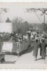 Outdoor view of homecoming parade, women seated on float with spectators walking down the street.