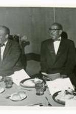 View of two men seated at a table with paper booklets.