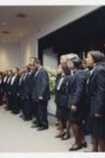Men and women, wearing suits with bow ties, sing in front of an audience at convocation.