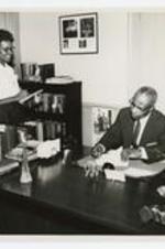 Interior view of George A. Sewell sitting at a desk with his secretary standing beside him.
