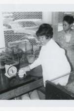 A young women holds a rat at a laboratory counter a scale and other science equipment in a classroom, as another woman looks on.
