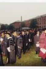 Men and women, wearing graduation caps and gowns, walk in a line between graduates in the audience at commencement.