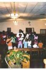 A congregation sings a hymn. Written on verso: Vacation Bible School - 2007. Carolyn Christian - Worship leader singing with children and other adults inside chapel.