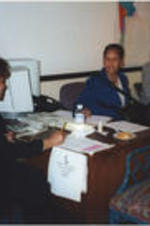 Two women sit at a desk during a "Get Out The Vote" phone bank event. Joseph E. Lowery sits in the background.