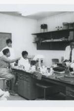 A man works with three young women at a laboratory table with trays and other science equipment in a classroom.