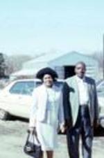 A couple dressed for church pose outside with cars and a barn in the background. Unknown location.
