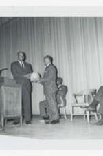 Two men stand with a basketball on stage at an event.