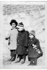 Three children stand in winter coats outside of a building