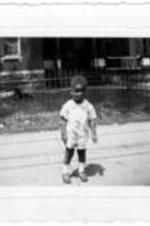 An unidentified little boy stands in front of an house on the sidewalk.