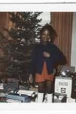 An unidentified girl in a blue sweater stands in front of a Christmas tree surrounded by presents.