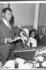 Dr. William Whatley (seated left) and others listen as Reverend Reginald Jackson speaks from the podium.