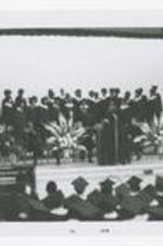 A group of men and women, wearing choir robes, stand on stage at commencement.