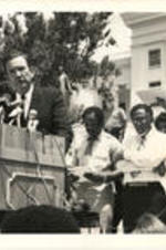 Ramsey Clark is shown speaking at a podium in front of John Nettles, Joseph and Evelyn Lowery, and others during an event in celebration of the 20th Anniversary of the Voting Rights Act. Written on verso: Former U.S. Attorney General Ramsey Clark speaks at the 20th anniversary ceremony of the Voting Rights Act of 1965 on the steps of the Alabama State Capitol in Montgomery. (L to R) Clark; Rev. John Nettles, Alabama SCLC President; Dr. Joseph E. Lowery, SCLC President; Mrs. Evelyn Lowery; National Convener of SCLC/WOMEN.