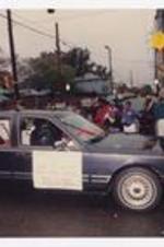 A young boy waves from the back window of a black car with sign "Women's Resource Center, Little Mr. and Miss Clark Atlanta University, Jasmine N. Mosley, Jonathan Wise Plummer, 1993-1994" in the homecoming parade.