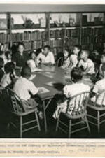 Hallie B. Brooks reads to students seated around a table. Written on recto: Story hour in the library of Oglethorpe Elementary School Library. Mrs. Hallie B. Brooks is the story-teller. [ca. 1940's]