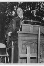 An unidentified man speaks from a pulpit at an outdoor commencement.
