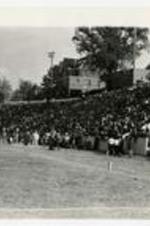 Outdoor view of people seated in stands with a marching band and buildings in the background.