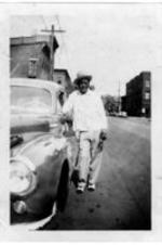 An unidentified man is leans on a car on a downtown street.