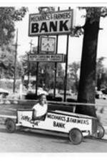 A young man (possibly John H. Wheeler' son) sits in a boxcar in front of a Mechanics and Farmers Bank. Painted on car, "Supecar".