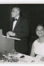 Rufus Clement speaks at a podium with a woman seated beside him.