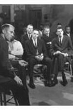 A group of men sit in chairs, watching a broadcast.