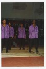 A group of men and women, wearing matching purple jackets, stand on the basketball court at a homecoming step show.