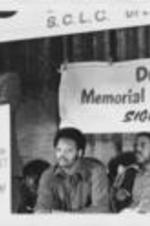 An unidentified speaker is shown speaking at a podium in front of Jesse Jackson and others during an Operation Breadbasket meeting.