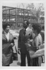 Two unidentified women speak with a man in a parking lot with a building under construction in the background.