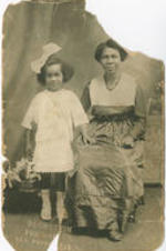 An unidentified woman sits in a chair while her daughter stands next to her holding a basket of lilies.