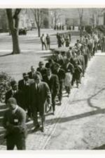 A goup of men and women walk down a brick sidewalk with college buildings in the background.
