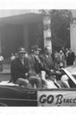 Bob Uecker, an unidentified man, and Ernie Johnson ride on the back of a parade car while Milo Hamilton sits in the passenger seat. Written on accompanying document: Broadcast Team, Bob Uecker-Milo-Ernie Johnson.