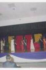 Young men and women, wearing formal wear with tiaras and sashes, stand in pairs on stage.