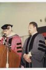 Thomas W. Cole, Jr. and Jesse Jackson, wearing graduation robes, stand at the podium at convocation.
