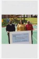 Dr. Walter Boradnax poses with another man and a woman holding a large check "Imiren Pharmaceuticals, Inc., September 2002, Pay to Clark Atlanta University $15,000," on football field.