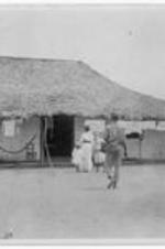People waking into a thatch roofed house in Liberia. Anna E. Hall wearing a white dress. Written on verso: visiting house of Mr. Mooney, native Commissioner.
