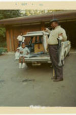 C. Eric Lincoln holds two large fish outside of a house, and a young girl sits on a station wagon tailgate and looks on.