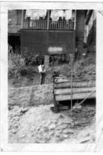 An unidentified holding a bag stands beneath a house on a hillside.