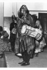 A man stands on a stage in an African toga carries a drum over his shoulder.